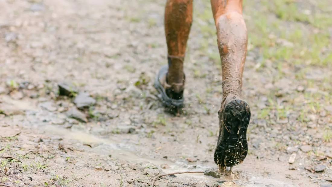 Gros plan sur les jambes et les chevilles d'un coureur en appui sur un sentier boueux et technique