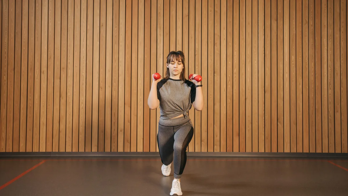 Femme réalisant une fente avec haltères en salle, un exercice de renforcement unilatéral sur une seule jambe pour coureur